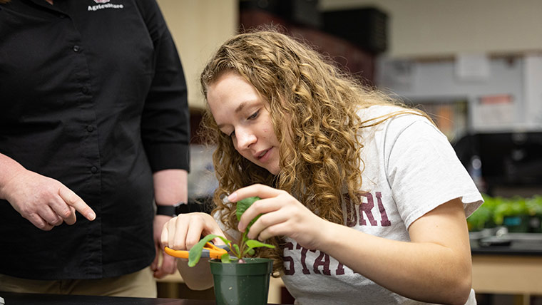 A student inspects a potted plant as a professor provides instruction.