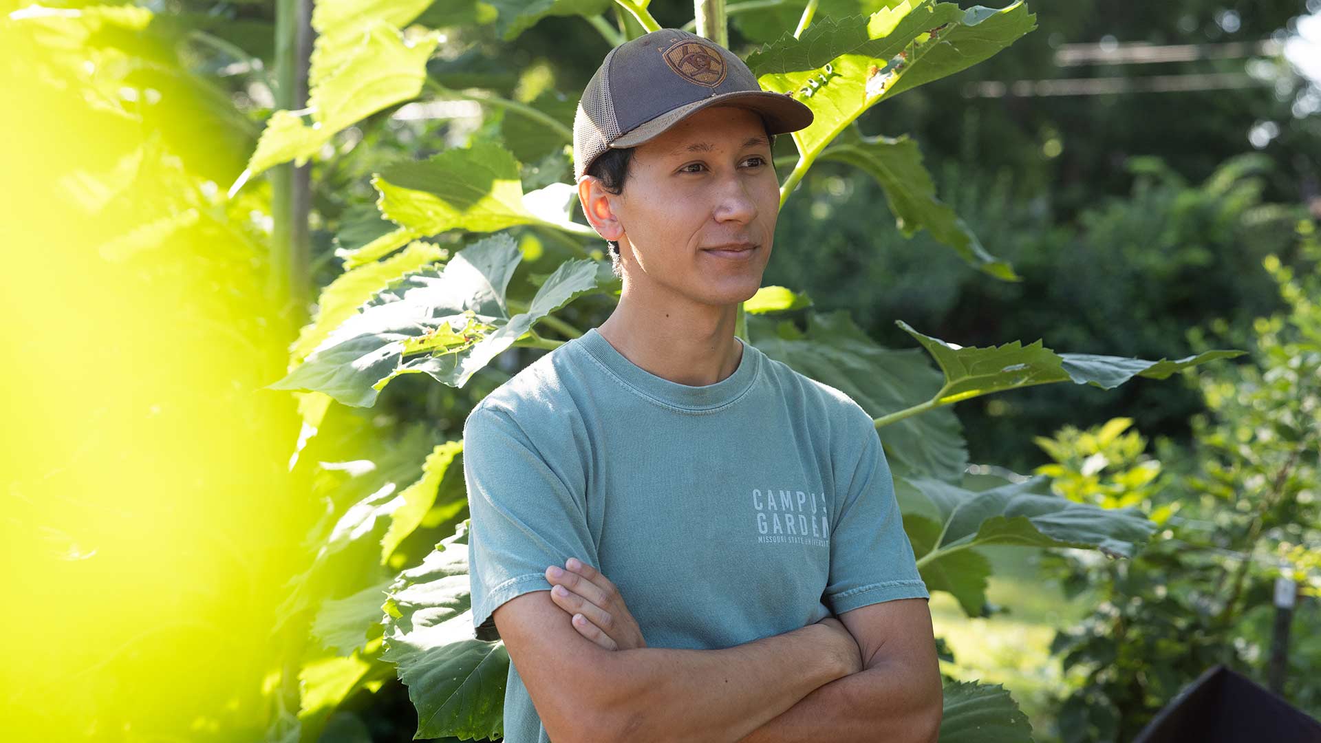 A person at peace while working in the campus garden. His arms are folded and he has a pleasant expression on his face.