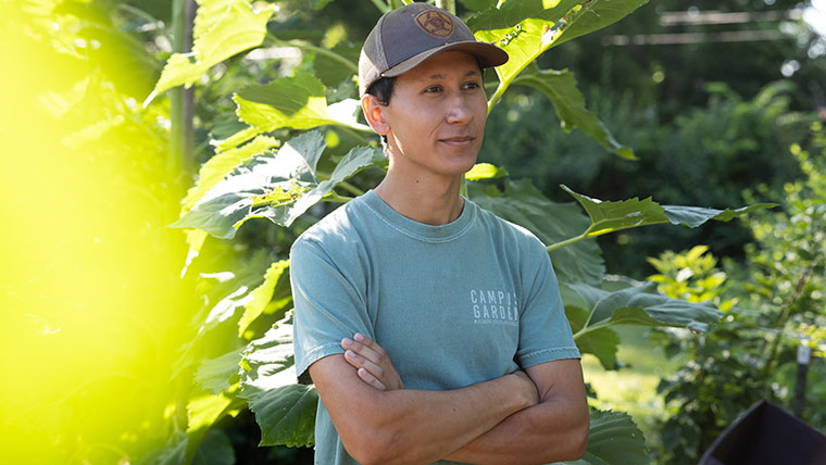 A person at peace while working in the campus garden. His arms are folded and he has a pleasant expression on his face.