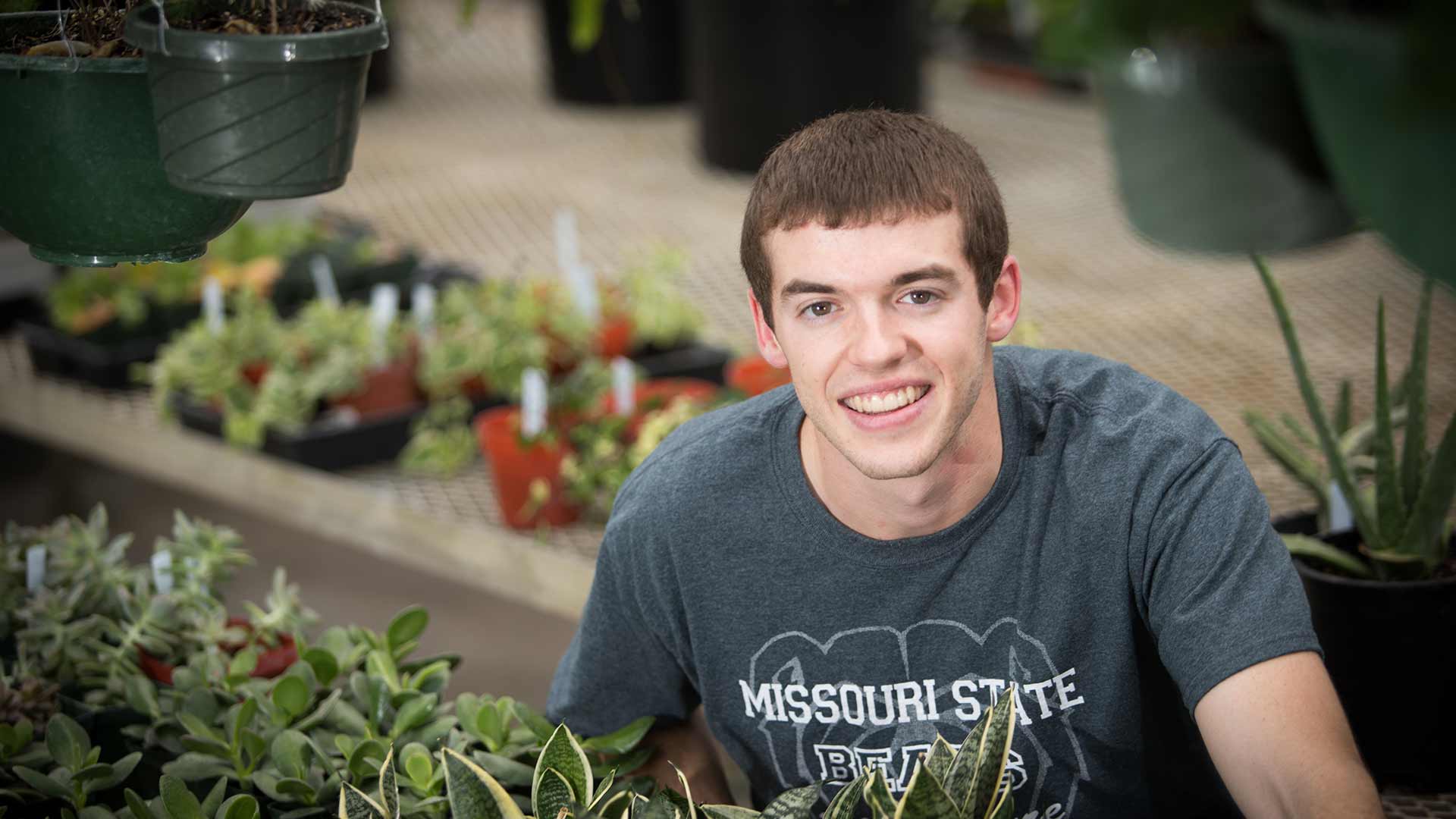 An agriculture student poses near rows of plants in a greenhouse.