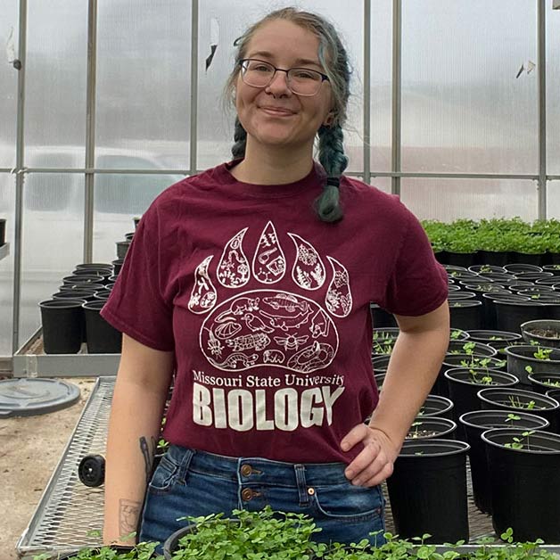 Abilene Mosher in a greenhouse wearing a maroon Missouri State biology t-shirt and blue jeans