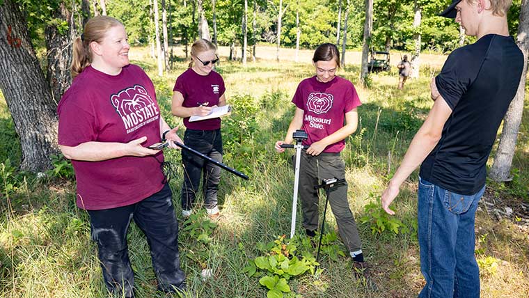 Dr. Melissa Bledsoe and three graduate students talk while conducting research in a forest area.