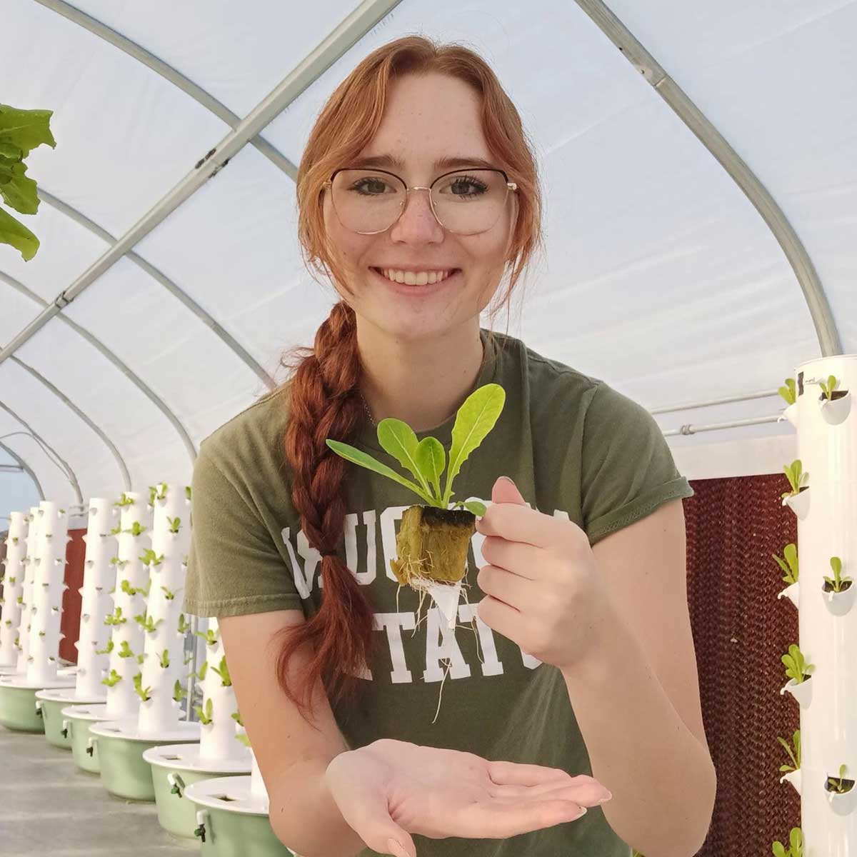 A Missouri State student in a greenhouse holds up a small plant while smiling.