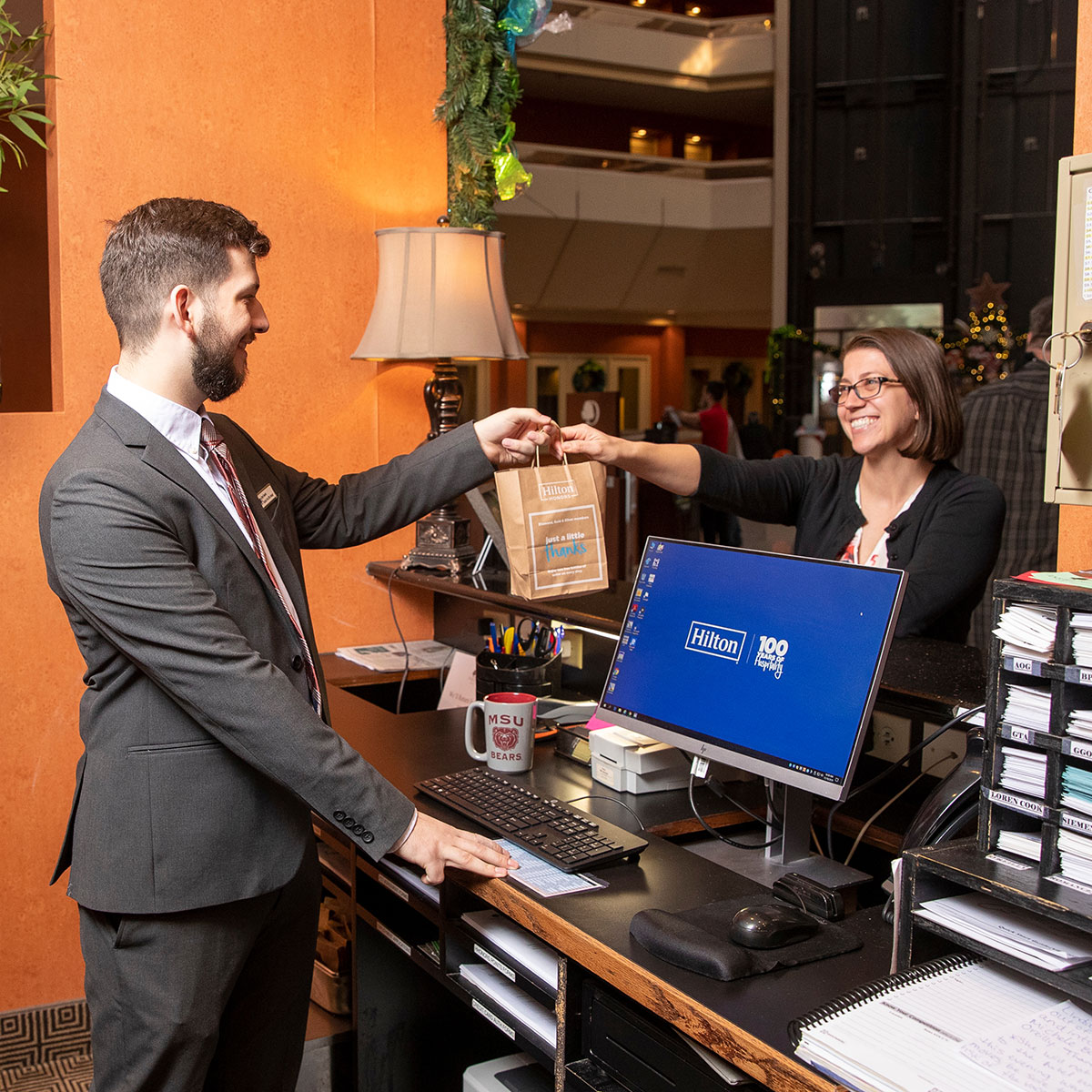 A student practices his hospitality skills while working as front desk staff at the Hilton Doubletree hotel. He hands a gift bag to a customer.